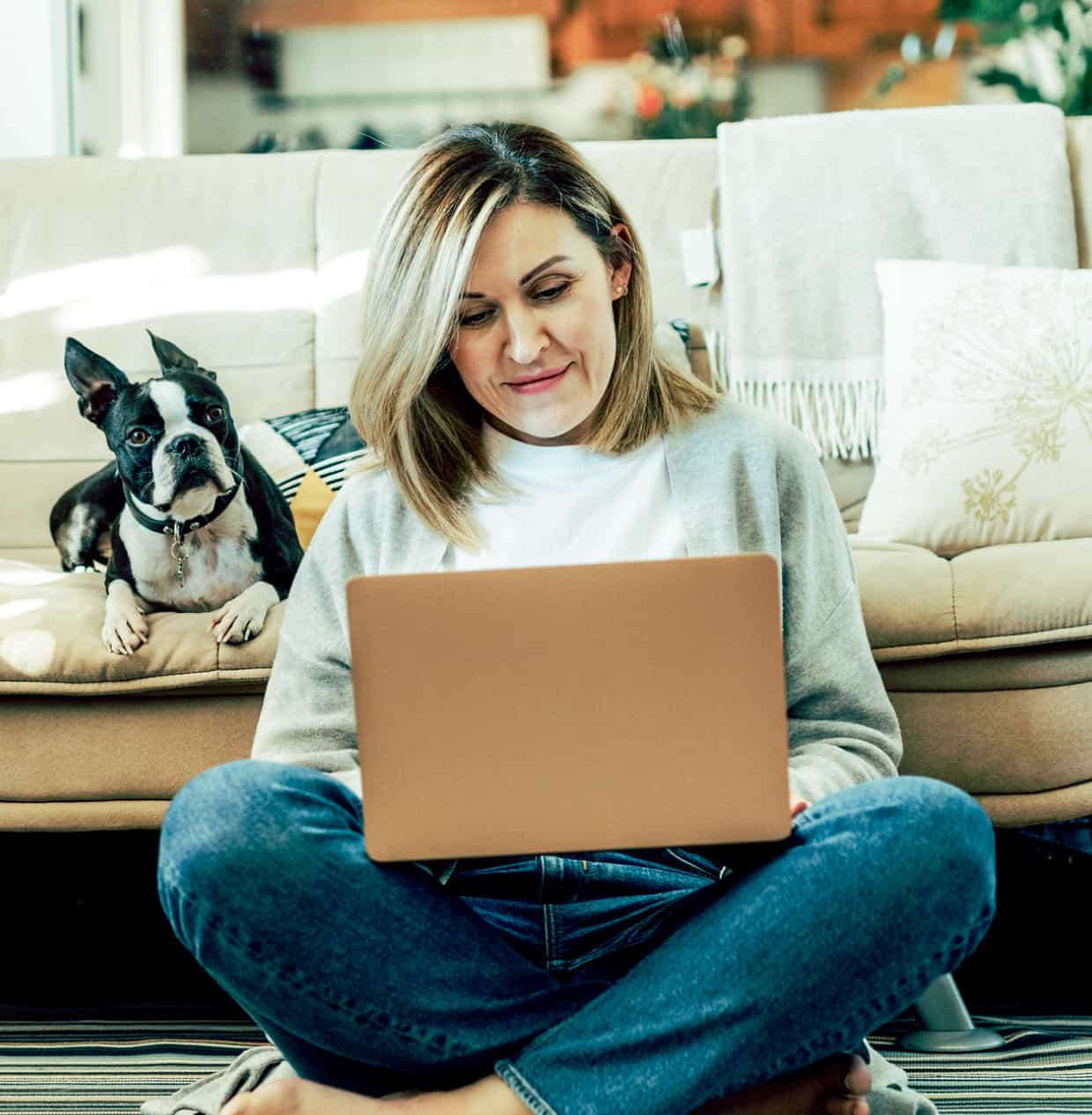 A woman sits cross-legged on the floor working on a laptop, with a dog on the couch behind her.