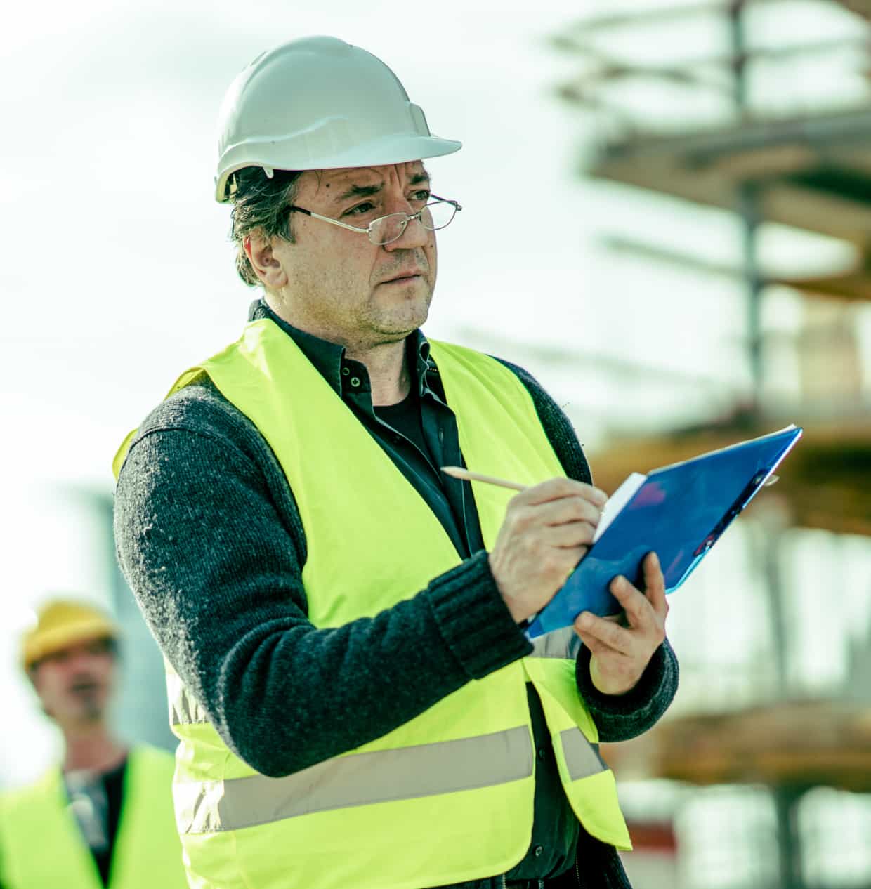 A man in a hard hat reviews information on a clipboard outdoors at a construction site.