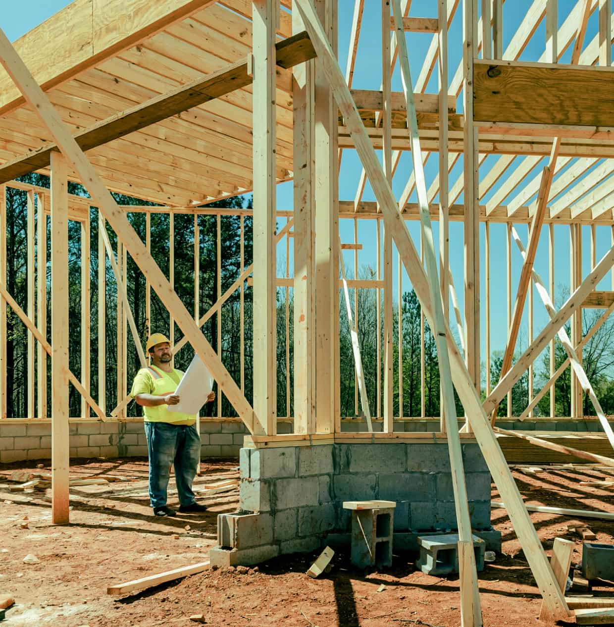 A construction worker in a reflective vest reviews plans in a partially framed building.