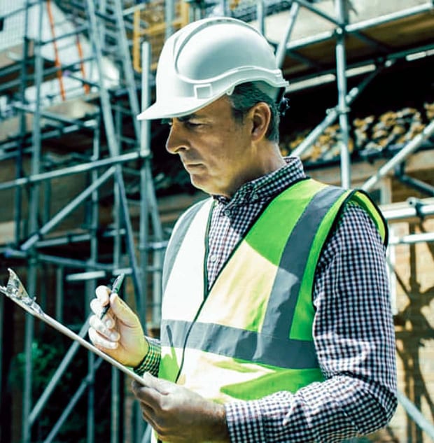 An inspector in a vest and hard hat reviews information on a clipboard at a project site.