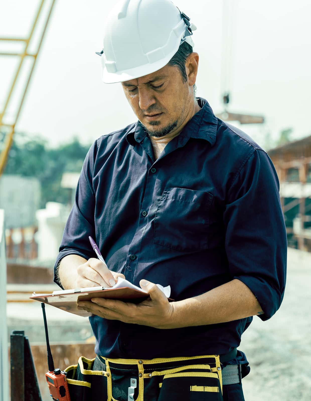A man in a hard hat writes something on a clipboard at a construction site.
