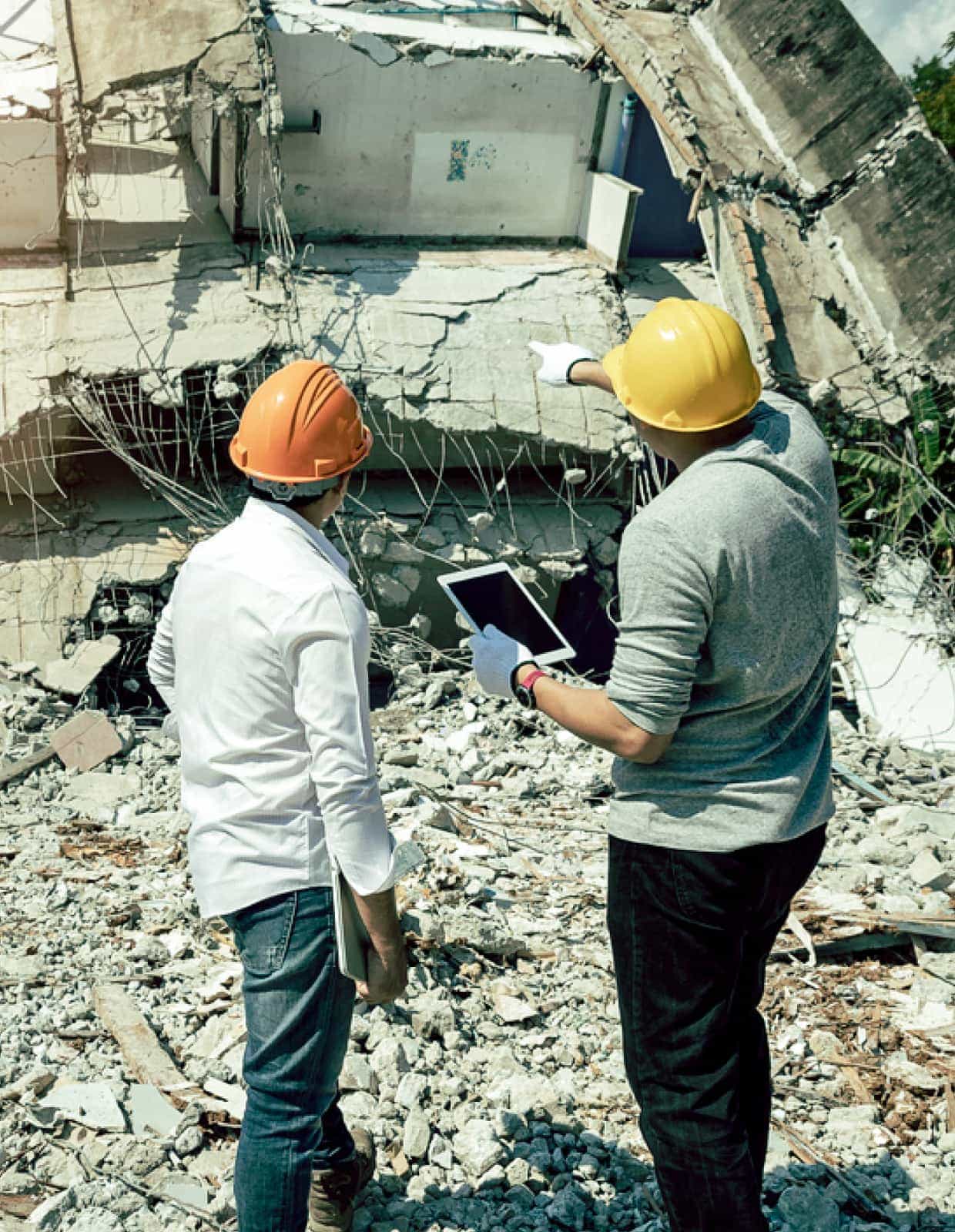 Two people in hard hats look at the rubble of a collapsed building.