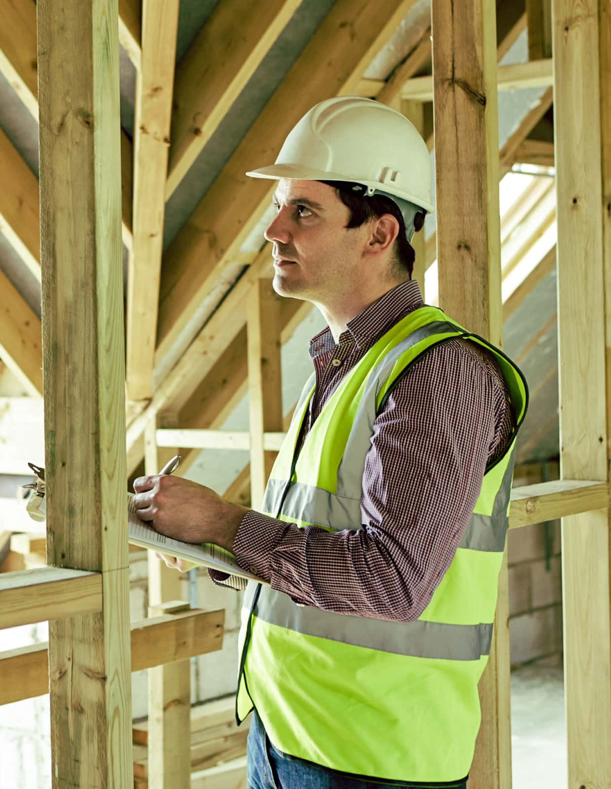 A construction worker in a framed building reviews information on a clipboard.