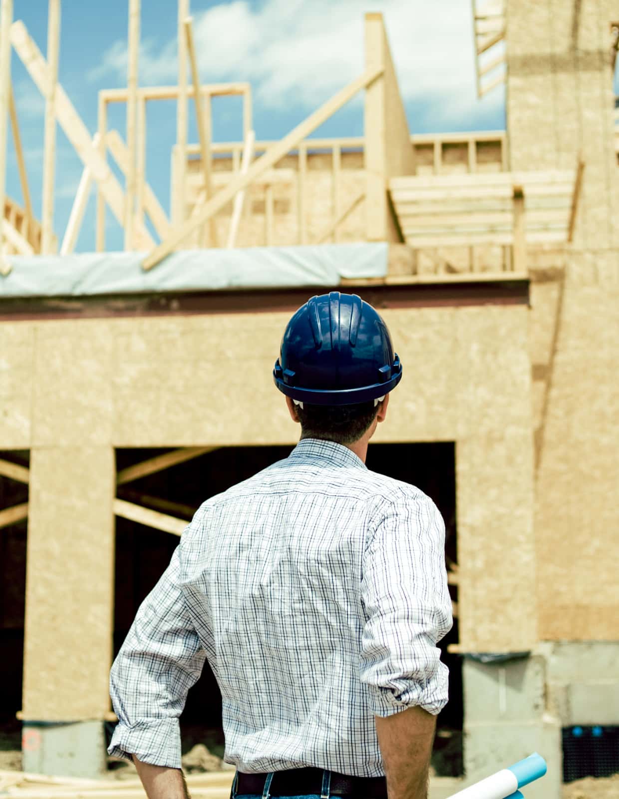 A man in a button-down shirt and a hard hat looks at an under-construction home.