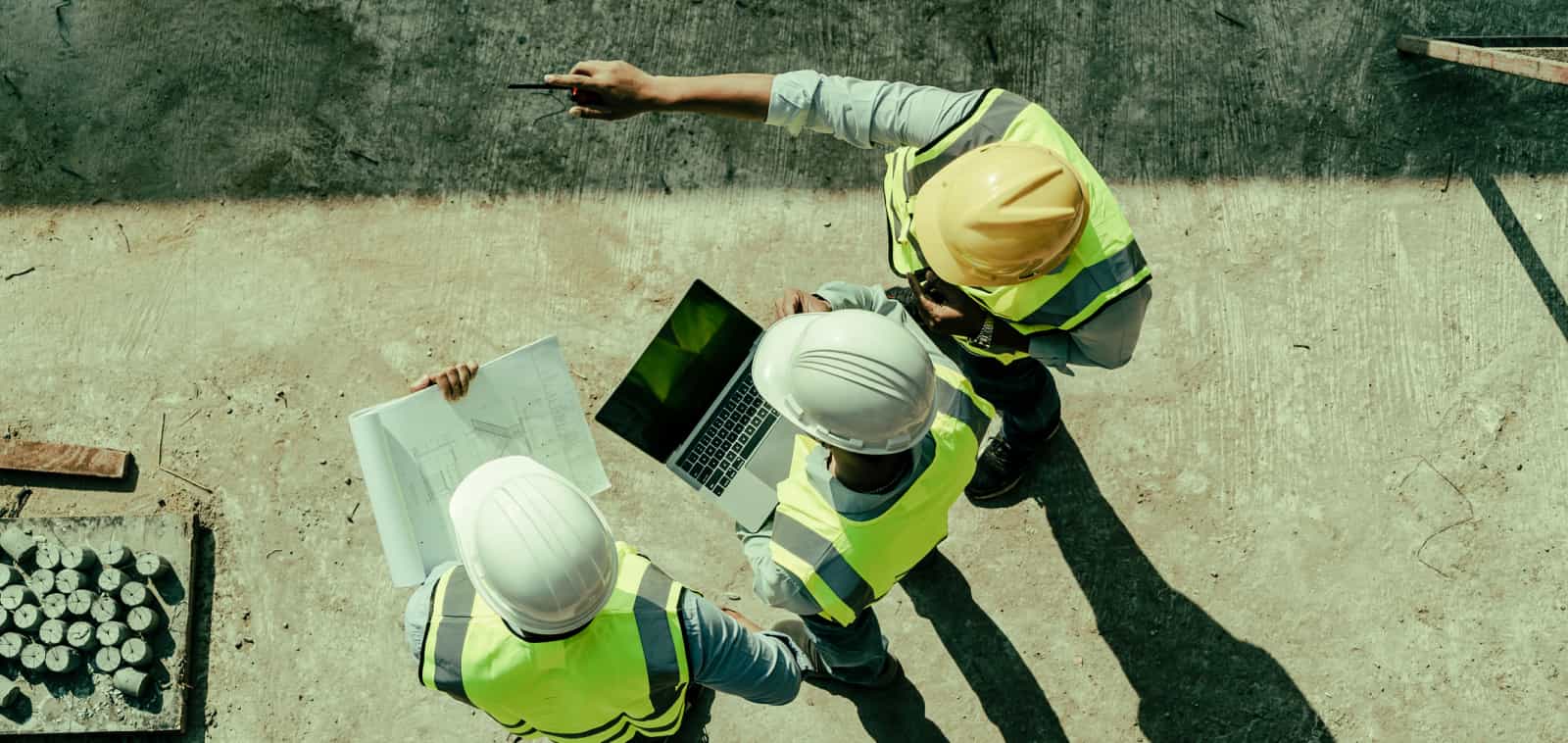 An overhead view of three people in hardhats reviewing paper plans and information on a laptop.