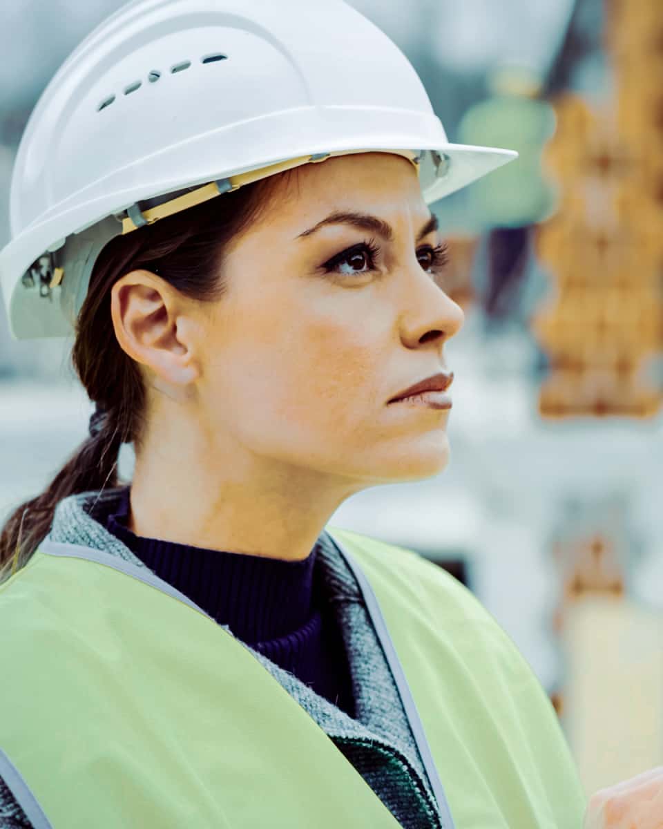 A close up of a woman in a hard hat looking up at a construction project.