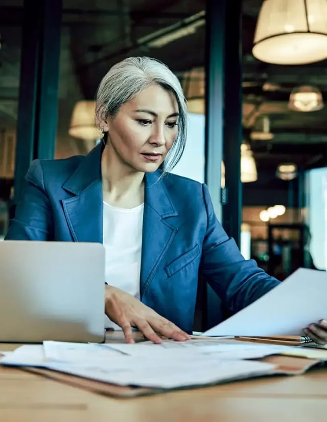 A woman in a blazer reviews papers on a desk next to her open laptop.